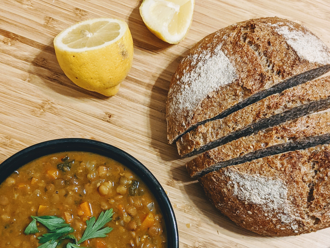 A bowl of lentil soup with a sliced loaf of bread and a lemon on a wooden surface