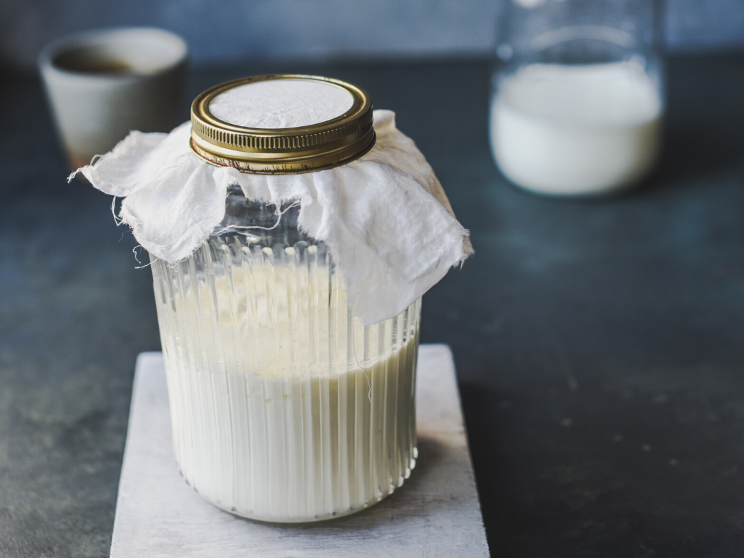 A jar of fermented milk with a cloth cover, on a surface with a glass of milk in the background.