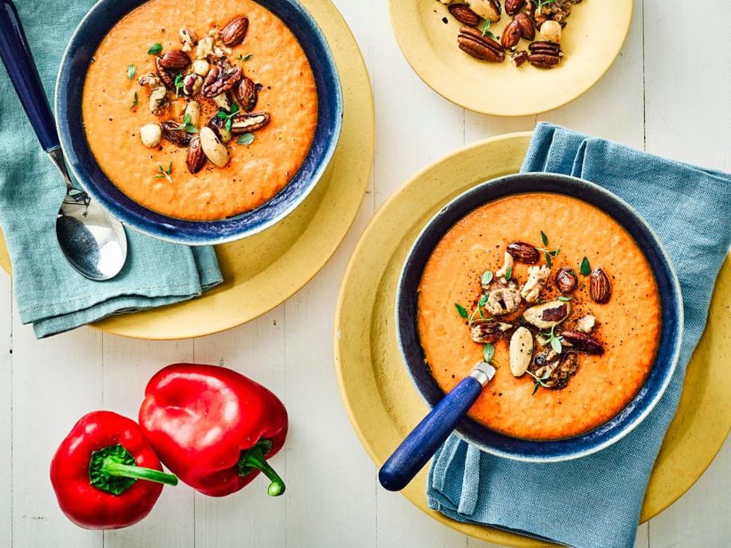 Two bowls of red lentil soup topped with nuts, with red bell peppers in the background.