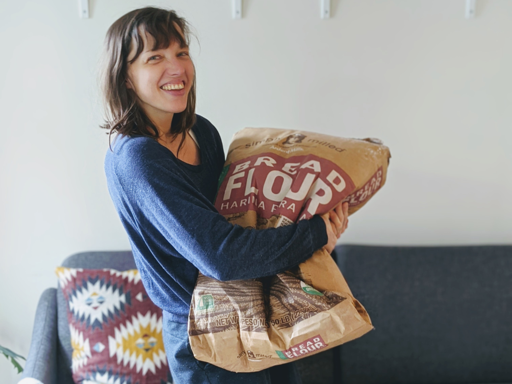 A woman smiling while holding a large bag of bread flour.