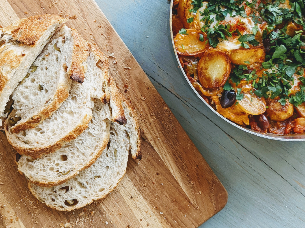 Sliced sourdough bread on a wooden cutting board next to a dish of baked potatoes and herbs.