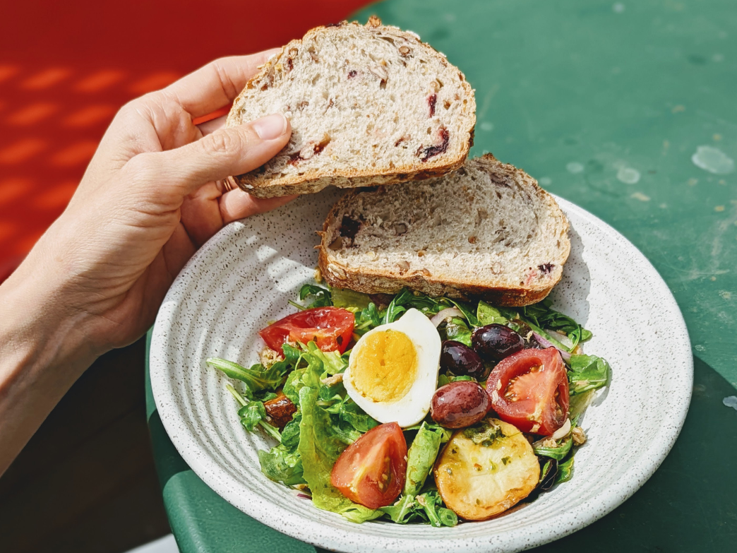 A hand holding two slices of whole grain bread above a bowl of salad with tomatoes, olives, and an egg.