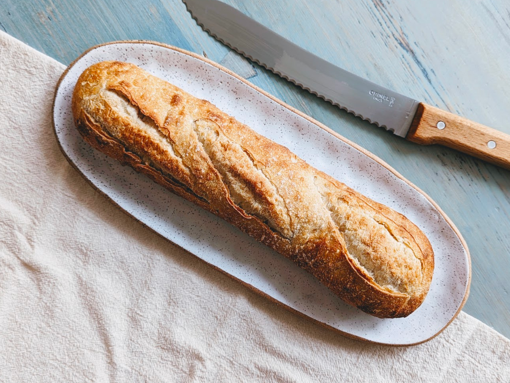 A freshly baked baguette on a plate with a serrated knife beside it.
