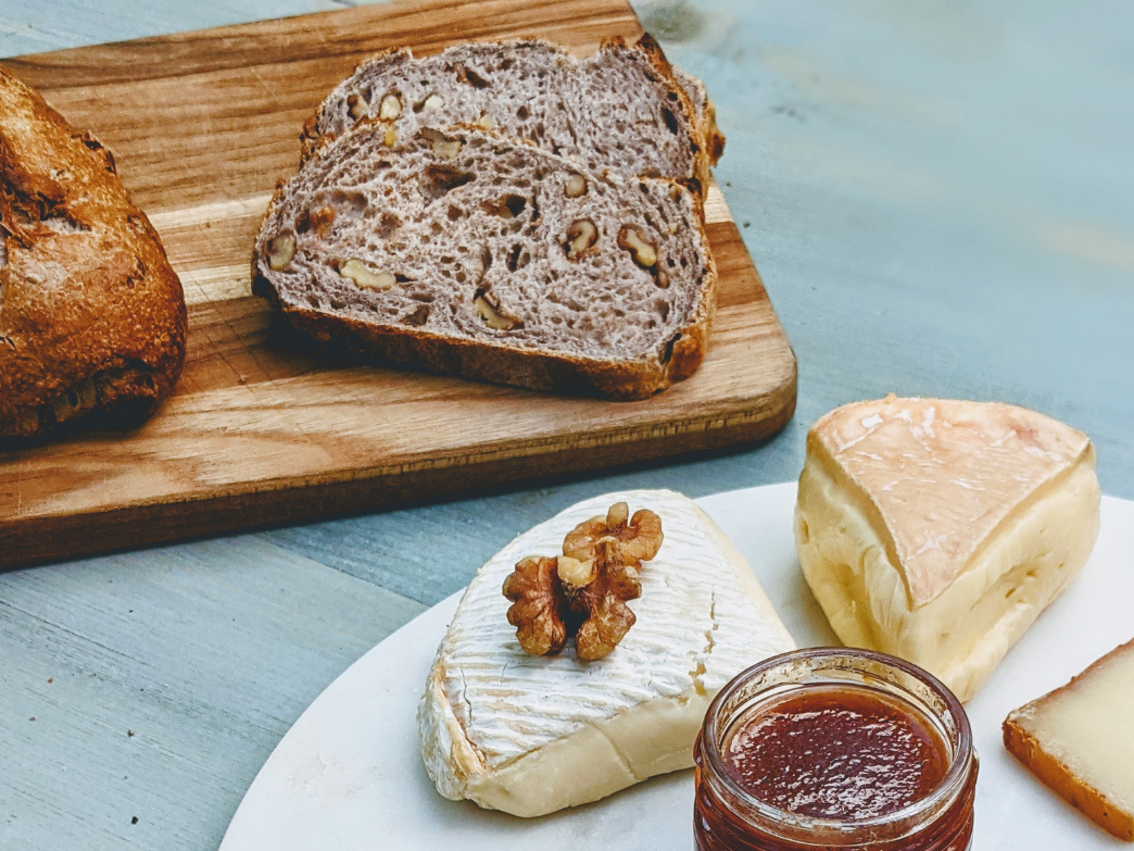 A wooden board with walnut bread, two types of cheese, and a jar of jam.