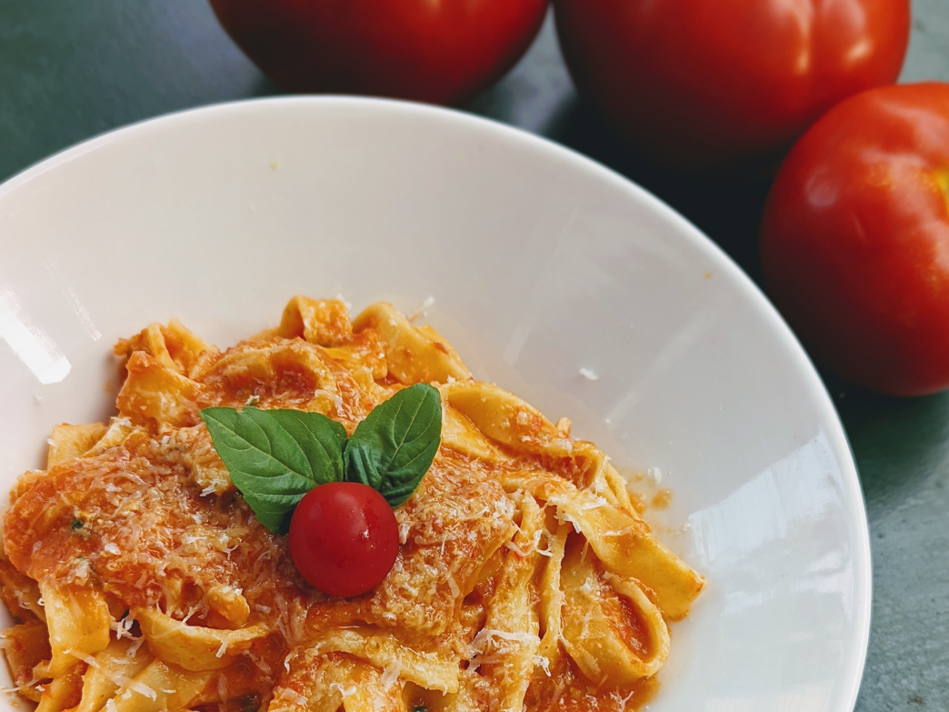 A bowl of fettuccine pasta topped with tomato sauce, cheese, a cherry tomato, and basil, with tomatoes in the background.