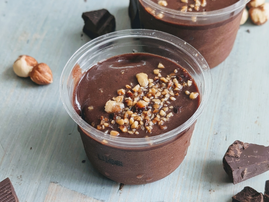 Close-up of a chocolate pudding cup showcasing glossy custard topped with chopped hazelnuts on pale blue wood