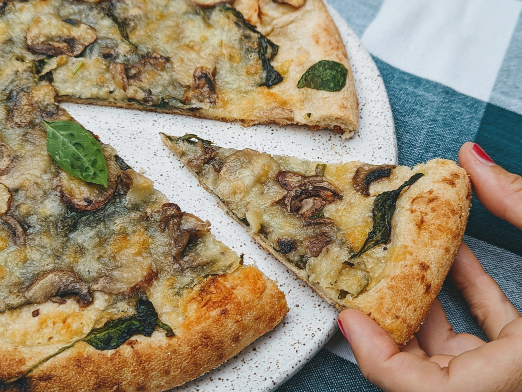 A hand reaching for a slice of mushroom and spinach pizza on a plate.