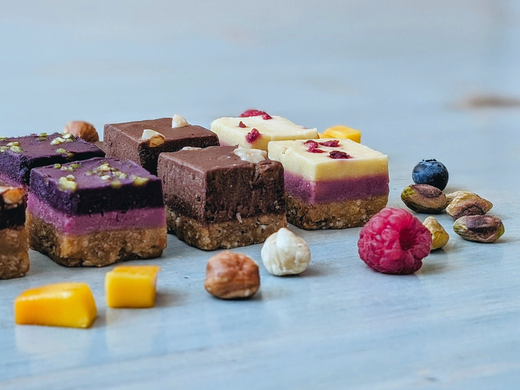 Close-up of layered dessert bars showing nut crust and purple berry, chocolate, and lemon layers on a light blue wooden surface