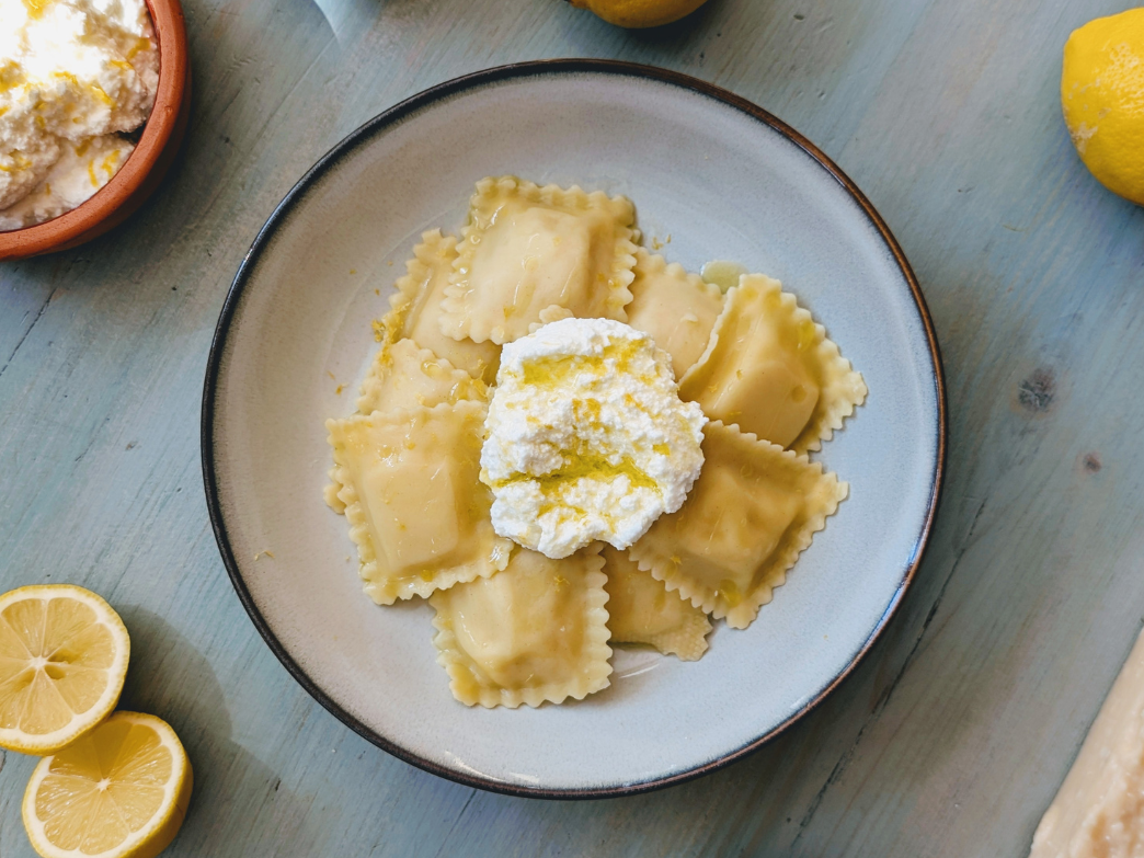 Plate of ravioli pasta topped with ricotta cheese and drizzled with olive oil, surrounded by lemon halves and a bowl of ricotta on a blue wooden surface.