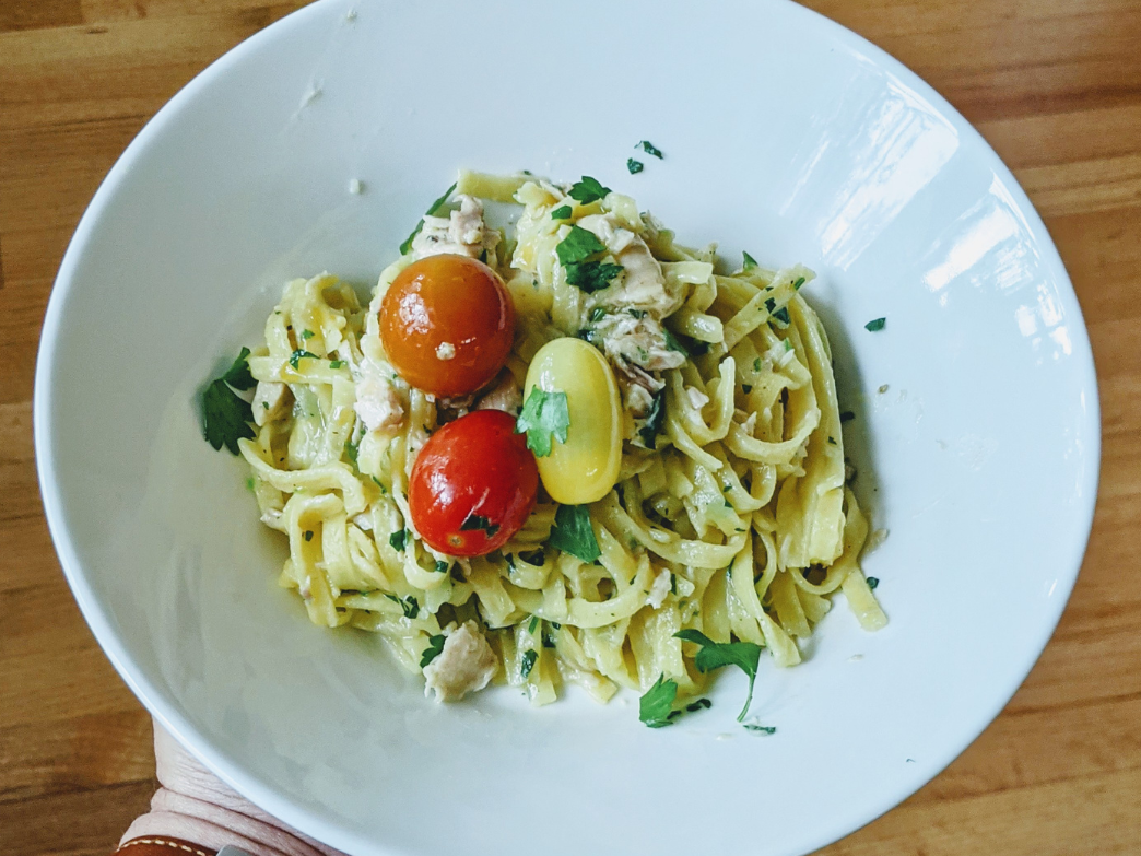 White bowl of plain egg fettuccine pasta with three cherry tomatoes and chopped parsley on a wood table