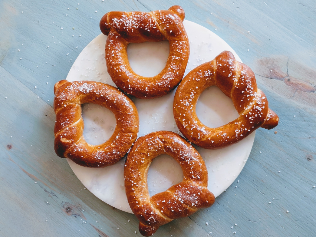 Four salted soft pretzels on a round white platter on a blue wooden table