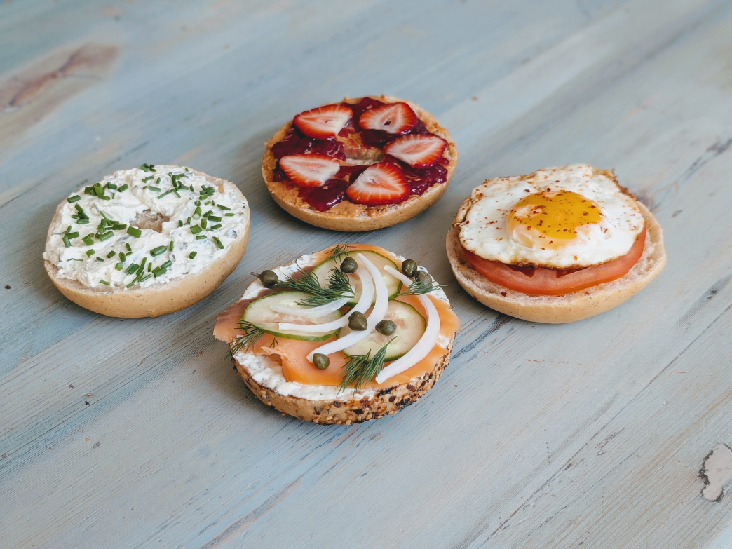 Four open bagels on blue wood: cream cheese, chives; jam, strawberries; fried egg, tomato; smoked salmon, onion, capers