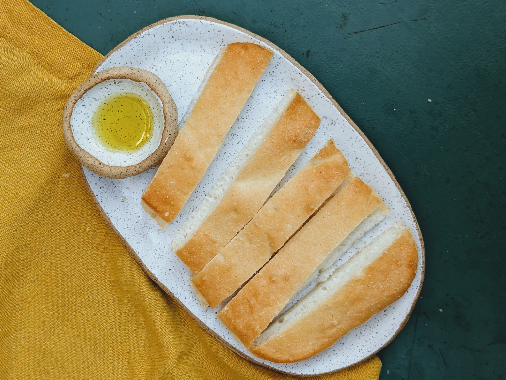 Sliced crusty bread arranged on a speckled oval plate with a small bowl of olive oil and a yellow cloth napkin.