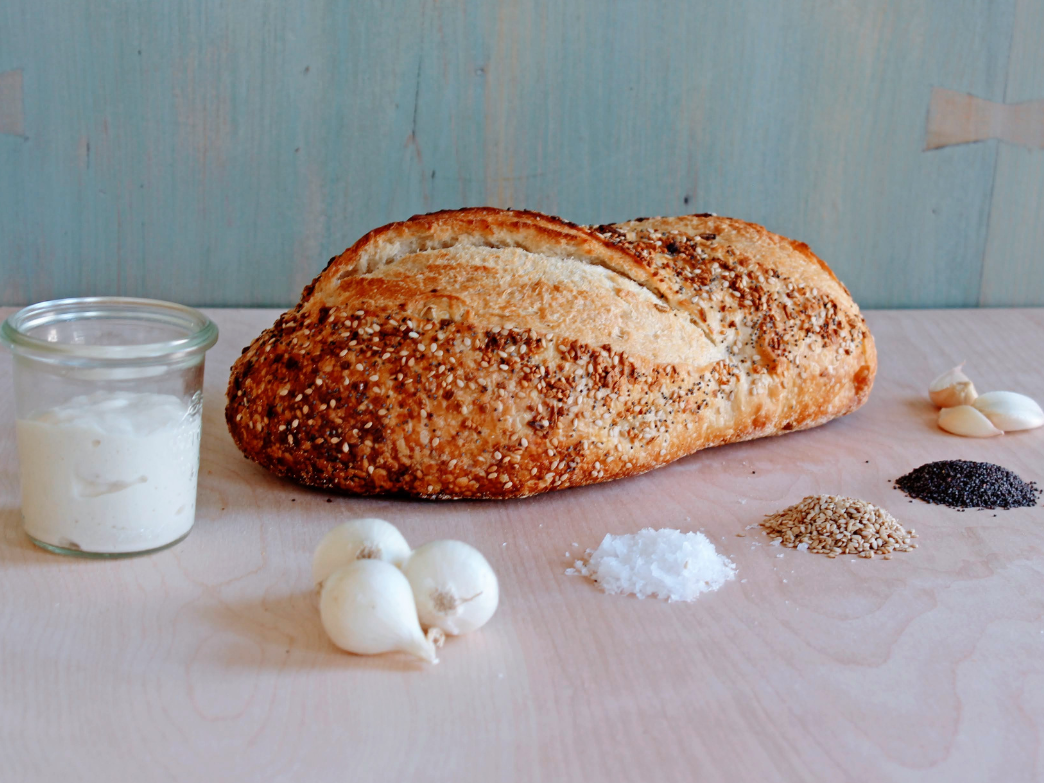 Seeded high-protein loaf on wooden board with jar of spread, garlic bulbs, salt and piles of seeds