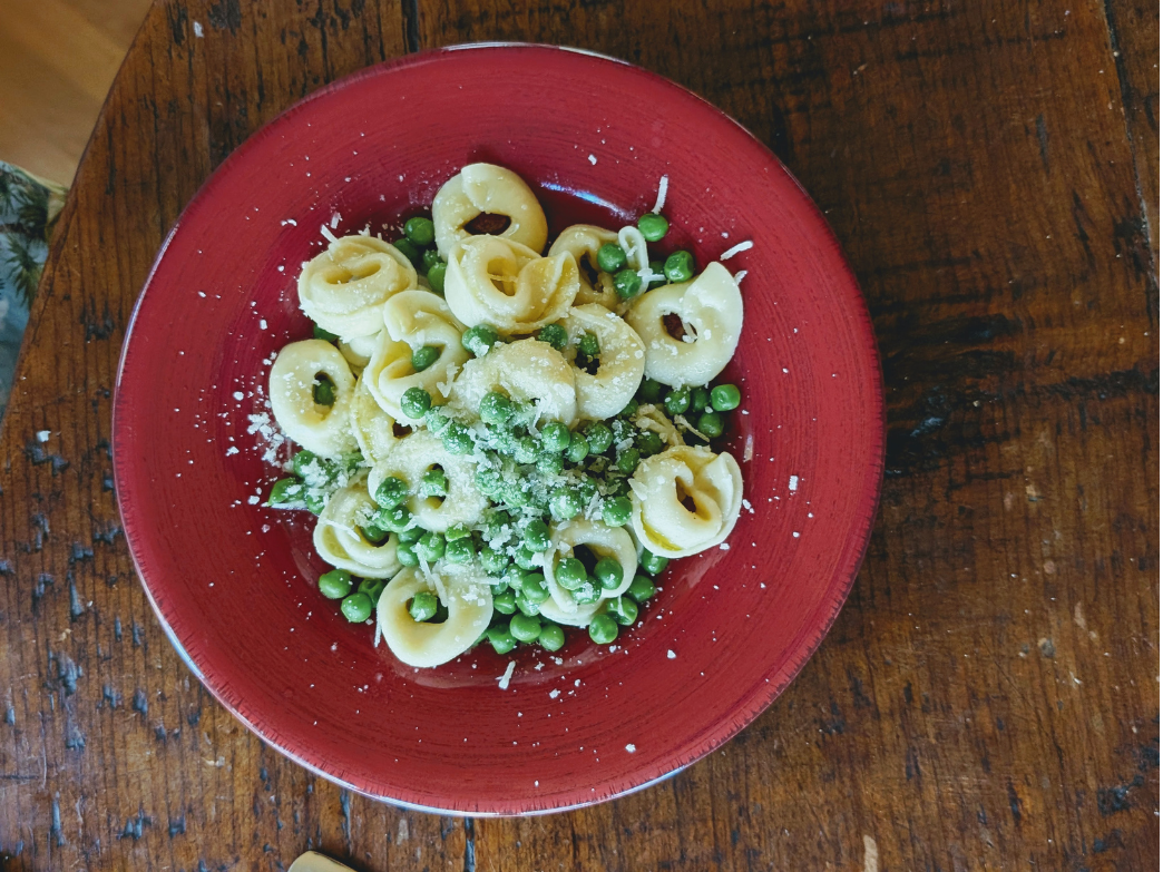 Red bowl of tortellini with peas and grated cheese on a wooden table.