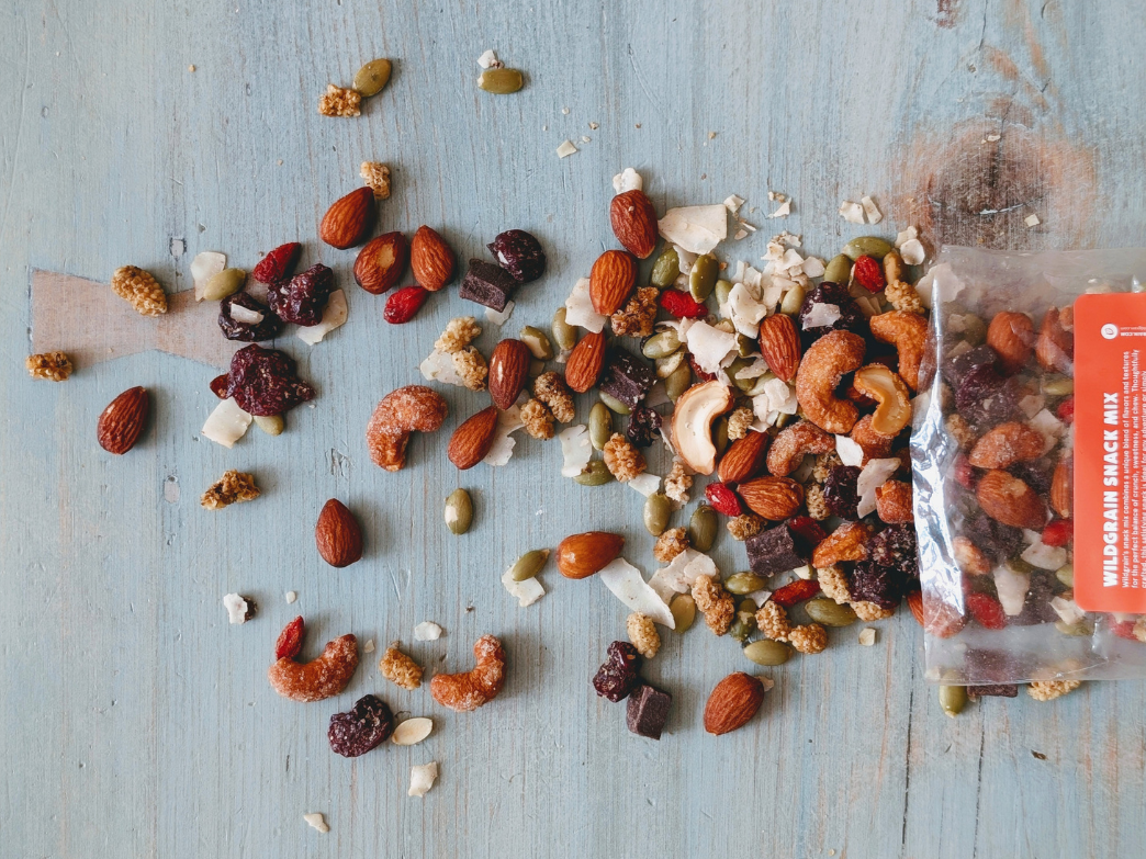 Mixed almonds, cashews, pepitas and dried fruit spilled from a clear bag labeled WILDGRAIN SNACK MIX on blue wooden table
