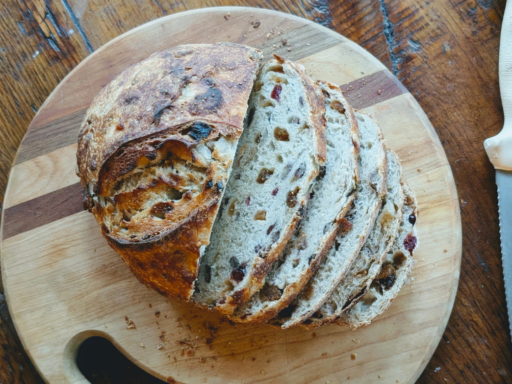 Sliced rustic sourdough loaf studded with dried fruit on a round wooden cutting board; serrated knife at right