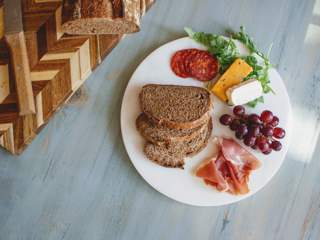 Three whole-grain bread slices on a plate with grapes, sliced cured meats, cheddar, brie, arugula, and a wooden cutting board.