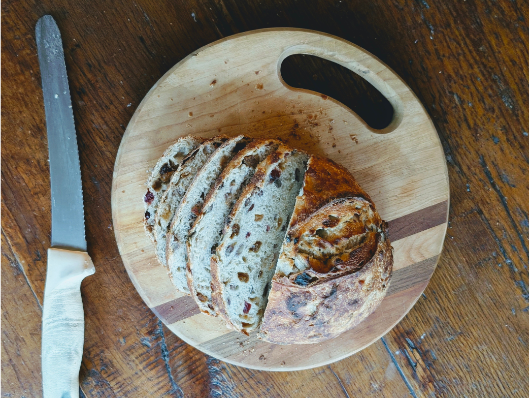 Sliced fruit-and-nut sourdough loaf on a round wooden cutting board beside a serrated knife