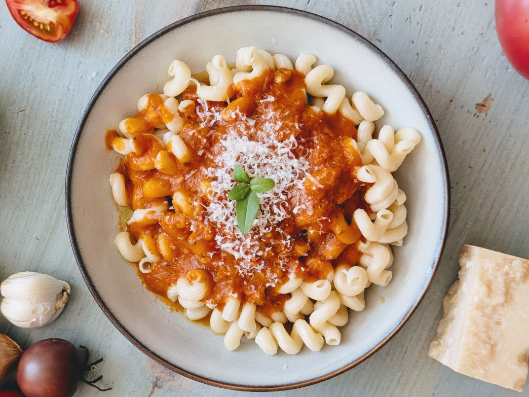 Bowl of spiral pasta topped with tomato sauce, grated Parmesan and a basil leaf, with garlic and tomato nearby