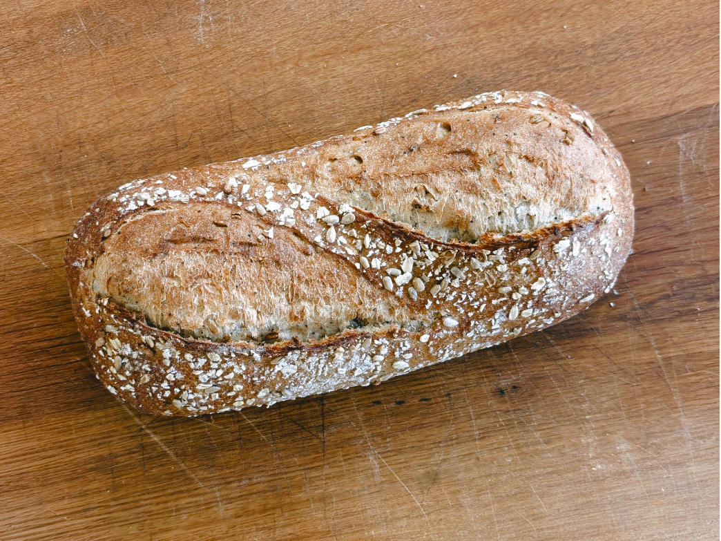Overhead view of whole-grain sourdough loaf showcasing golden crust, oats, and center scoring on wooden cutting board