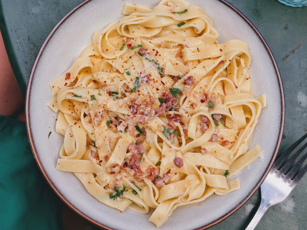 Plate of wide ribbon pasta with creamy sauce, crispy pancetta, and chopped chives on a green table