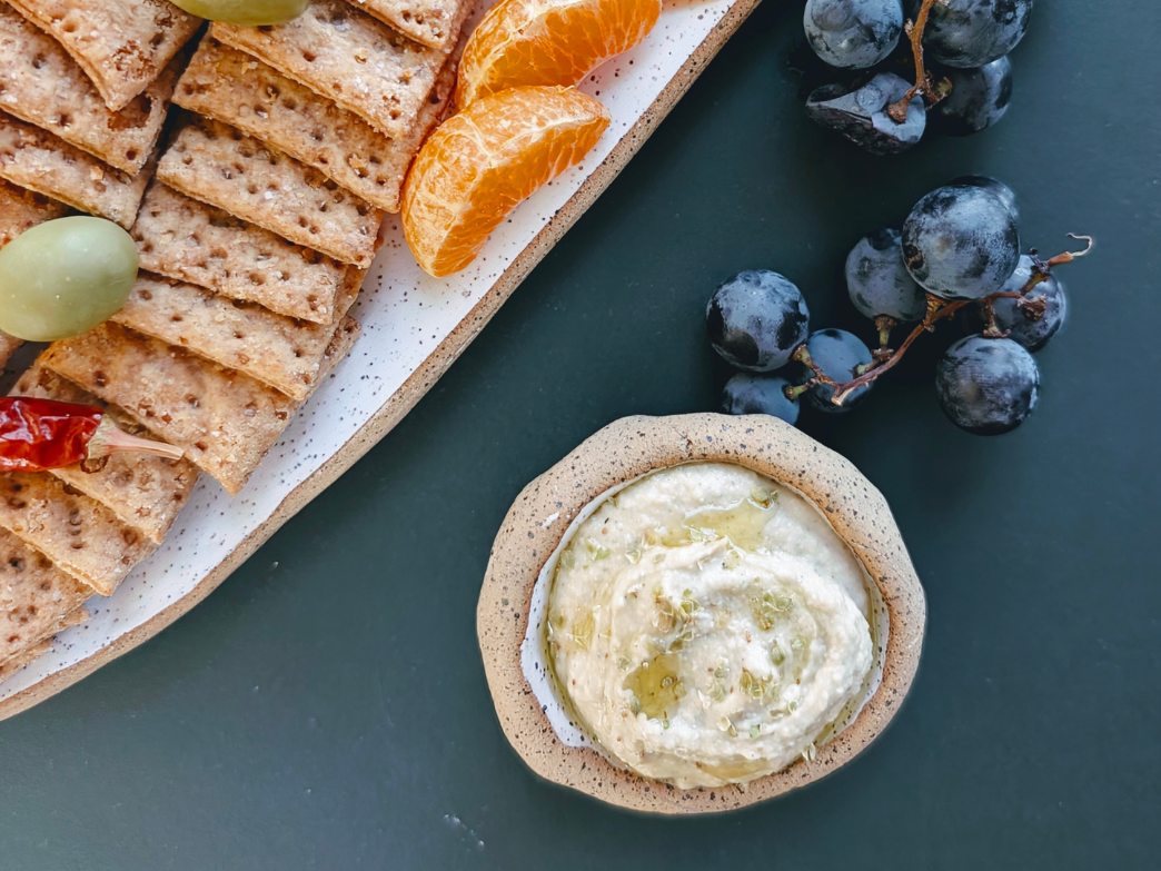 Bowl of hummus drizzled with olive oil beside whole-grain crackers, clementine slices, and black grapes on a dark background