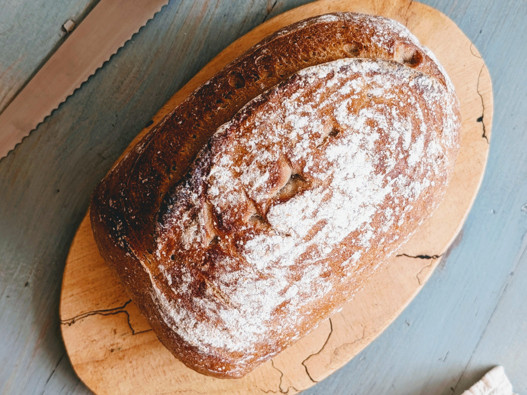 Angled top view of a whole-grain loaf dusted with flour showing crust and texture on a wood cutting board with a serrated knife