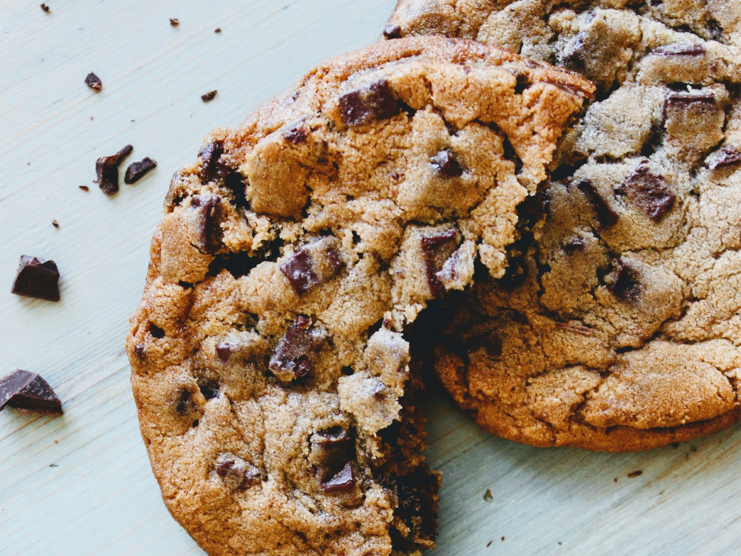 Two chocolate chip cookies, one broken, on a pale wooden tabletop with scattered chocolate chunks and crumbs