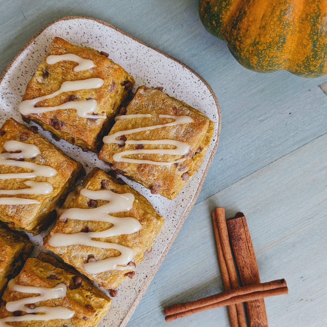 A plate of Pumpkin Cinnamon Biscuits drizzled with icing, surrounded by a pumpkin and cinnamon sticks.