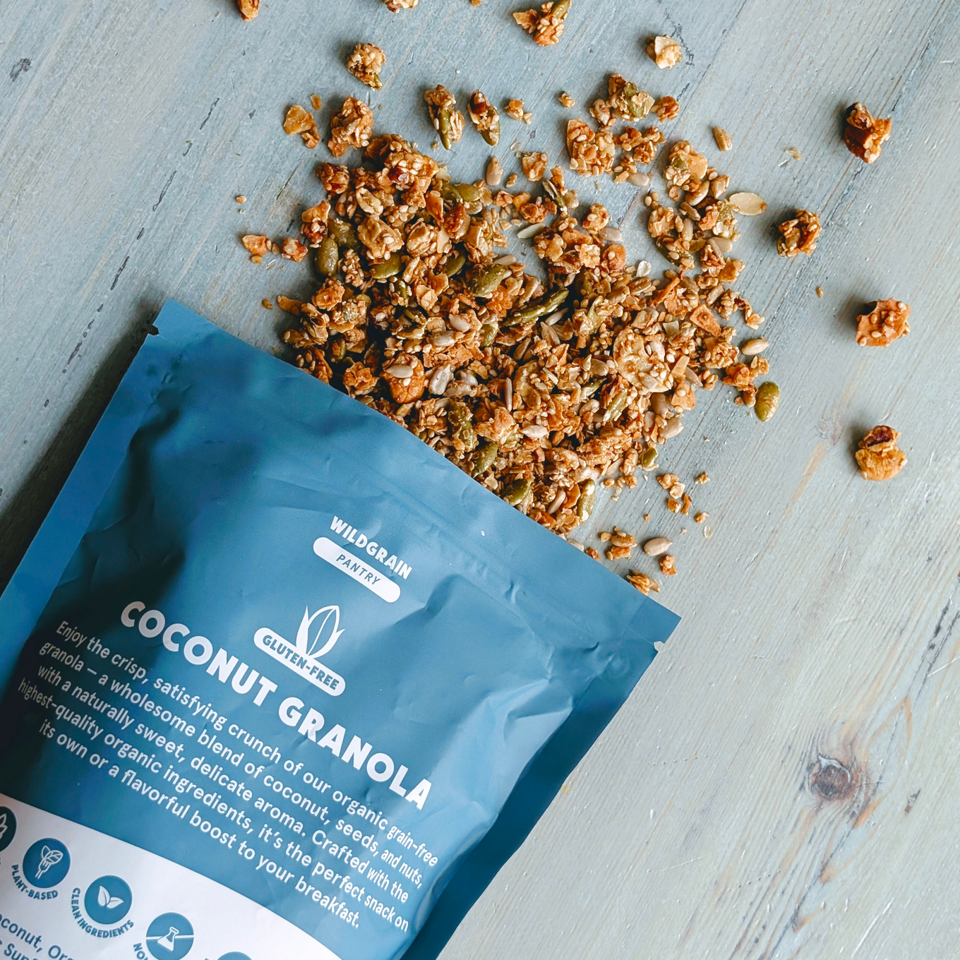 Top-down view of Coconut Granola pouch showcasing spilled granola and gluten-free badge on light wood background.