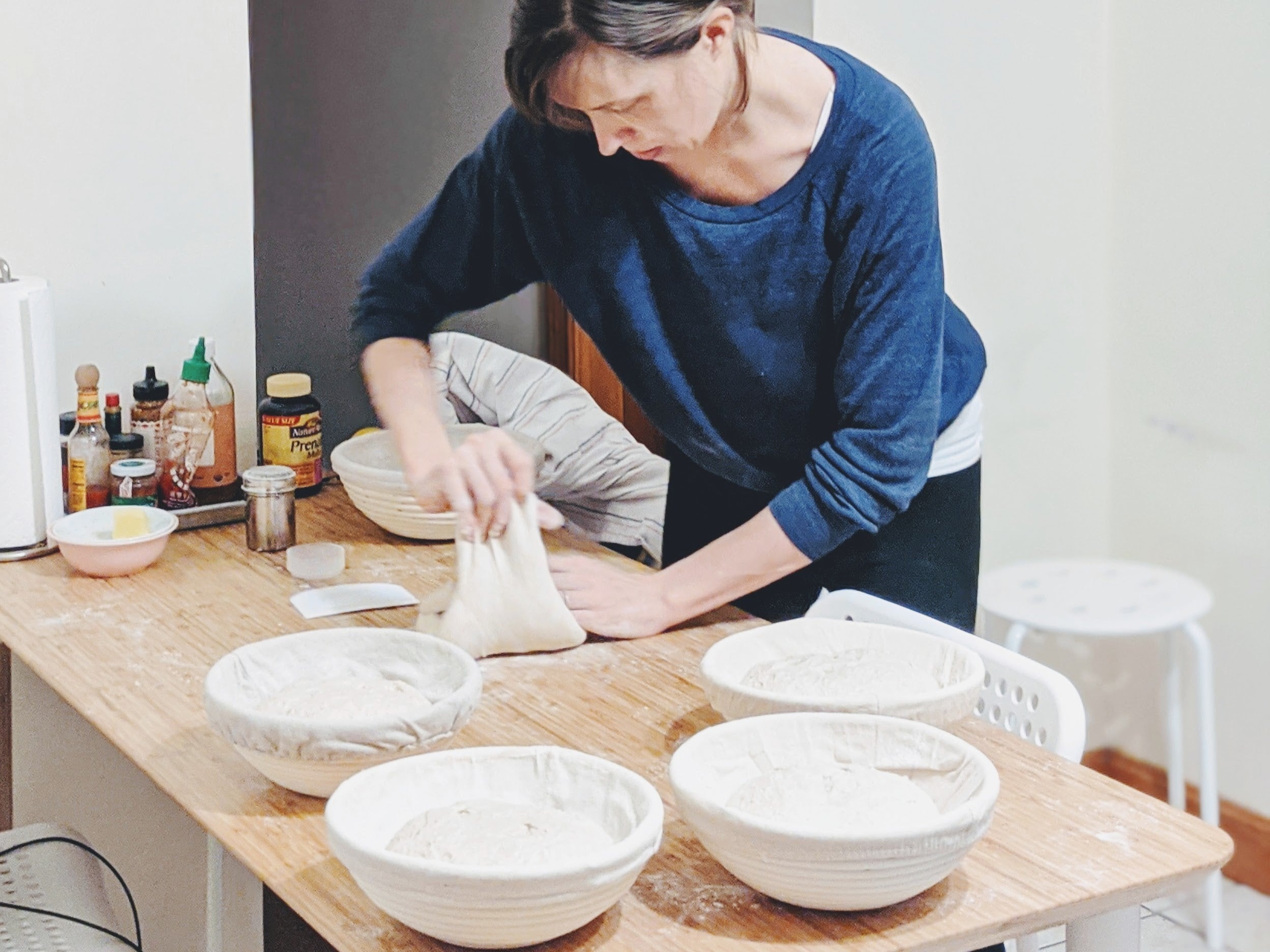 A woman kneading dough in a kitchen with bowls of dough on the table.