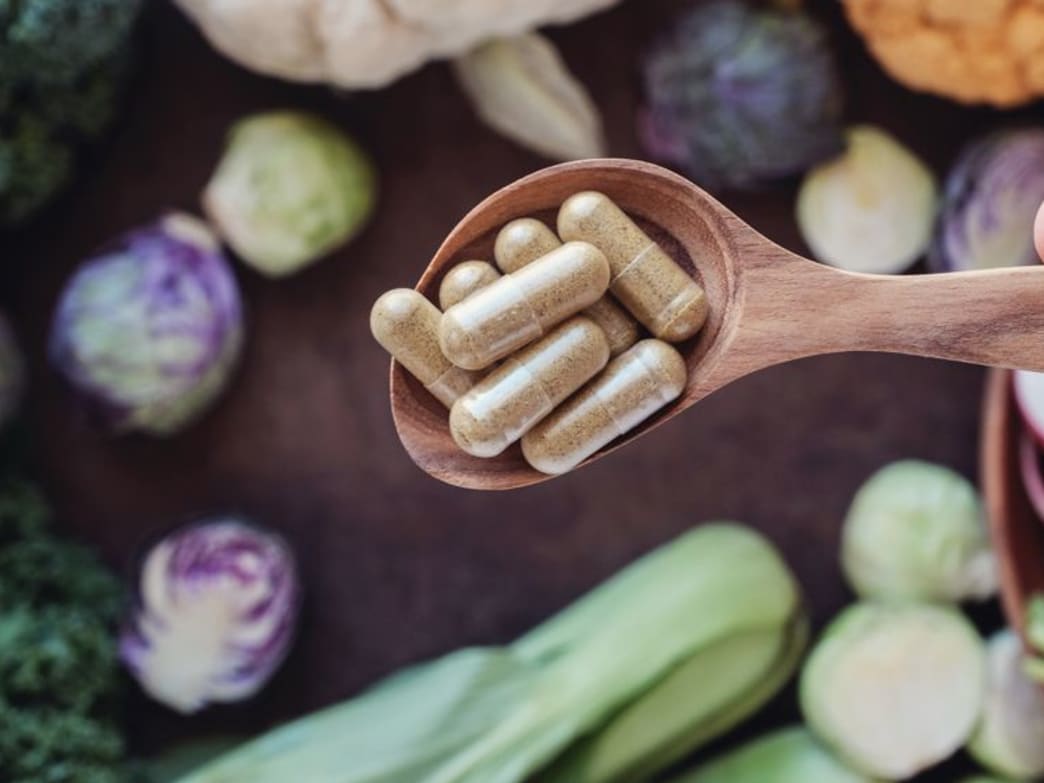 A wooden spoon holding dietary supplements surrounded by fresh vegetables.