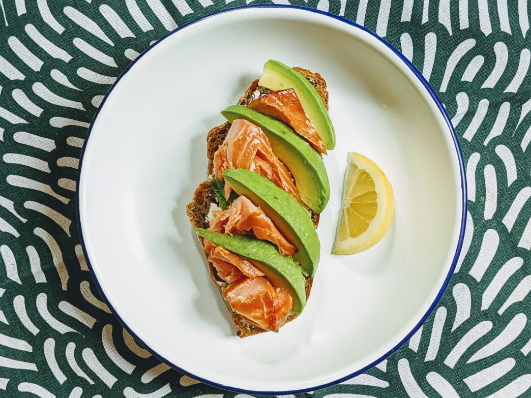 Open-faced salmon toast with avocado slices and a lemon wedge on a patterned plate