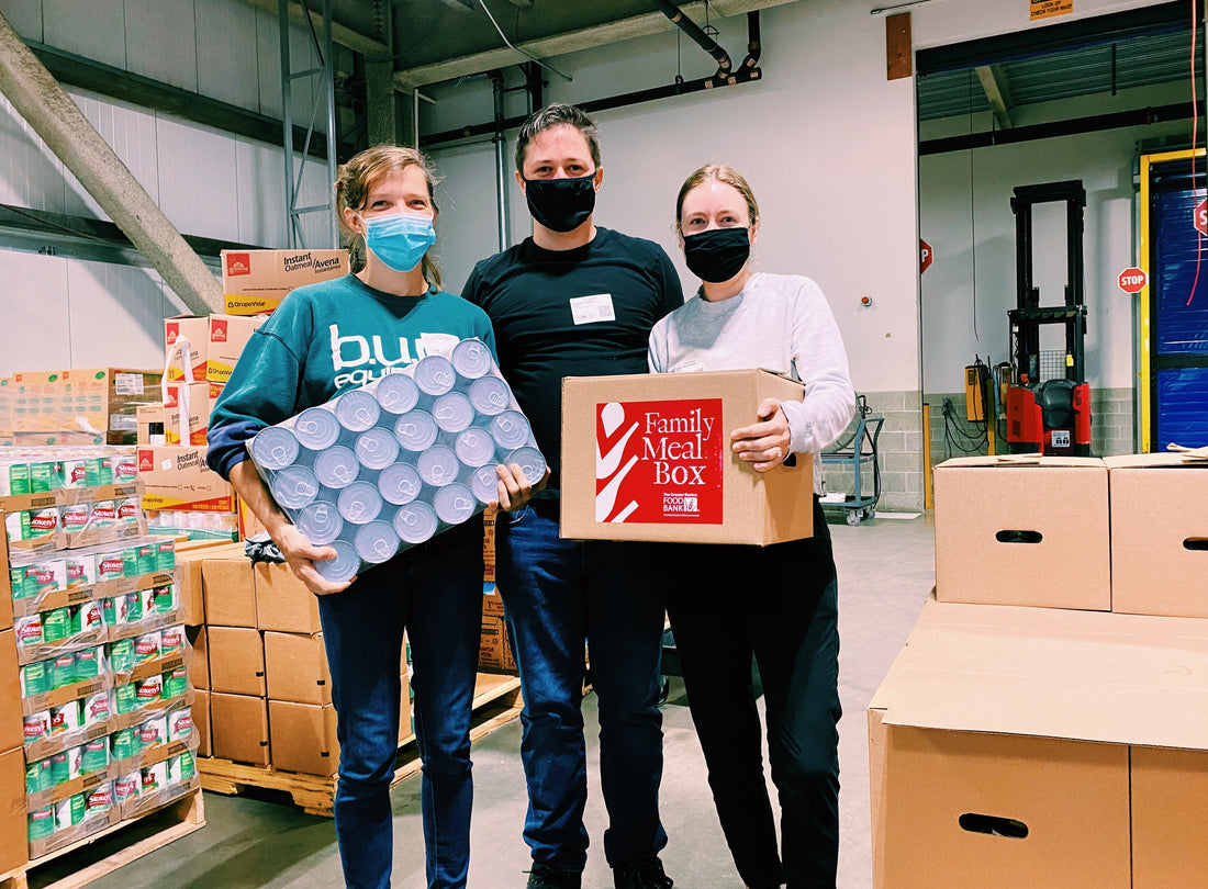 Three masked volunteers holding cans and a box labeled 'Family Meal Box'; nearby boxes say 'Instant Oatmeal/Avena' and 'STOP'