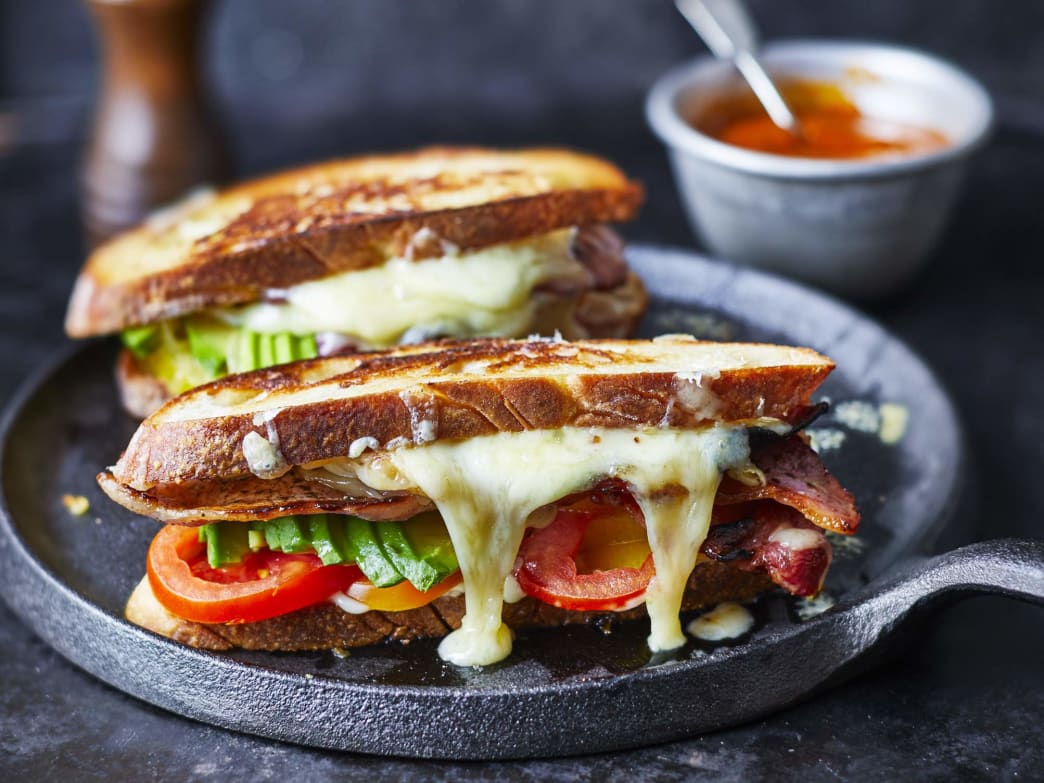 Two grilled cheese sandwiches with melted cheese, bacon, and vegetables on a cast iron skillet