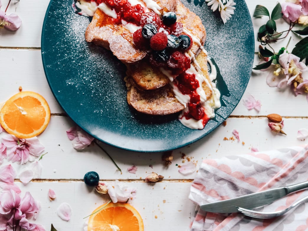 Plate of French toast topped with berries and sauce, surrounded by flowers and orange slices.