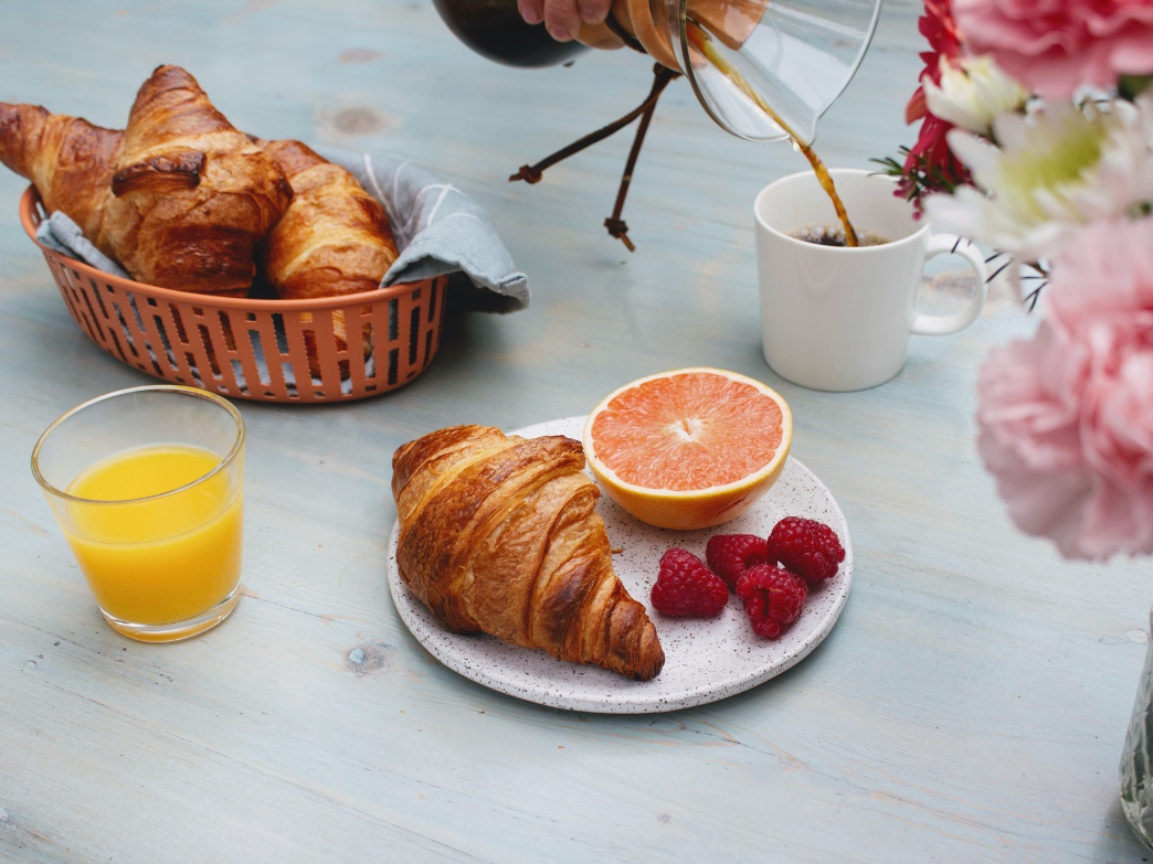 A plate with a croissant, raspberries, and half a grapefruit, with a glass of orange juice and coffee being poured.