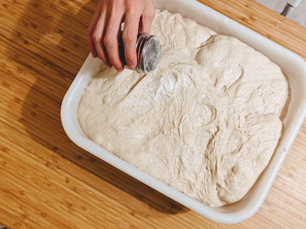 A hand sprinkling flour over a bowl of dough on a wooden surface.