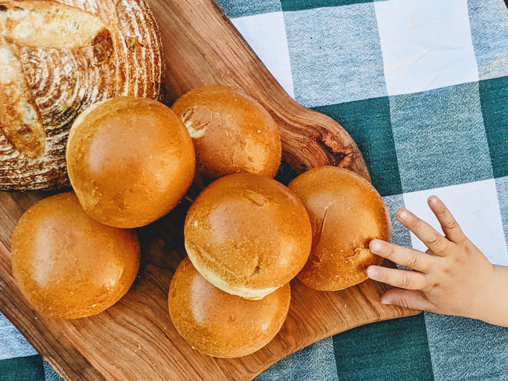 A wooden board with several golden brioche rolls and a child's hand reaching for one.
