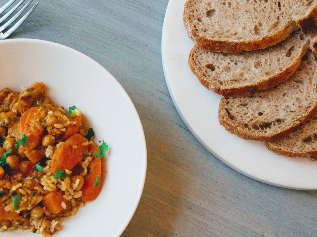 A plate of sliced sourdough bread next to a bowl of stew with vegetables and grains.