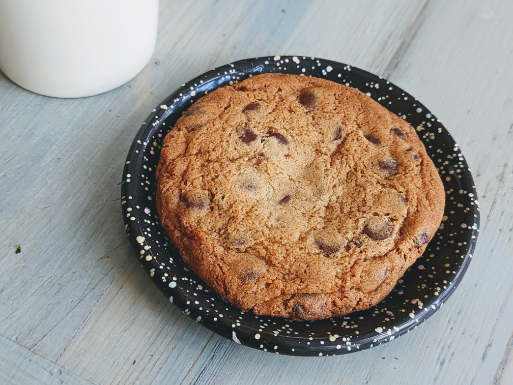 A large chocolate chip cookie on a speckled plate next to a glass of milk.