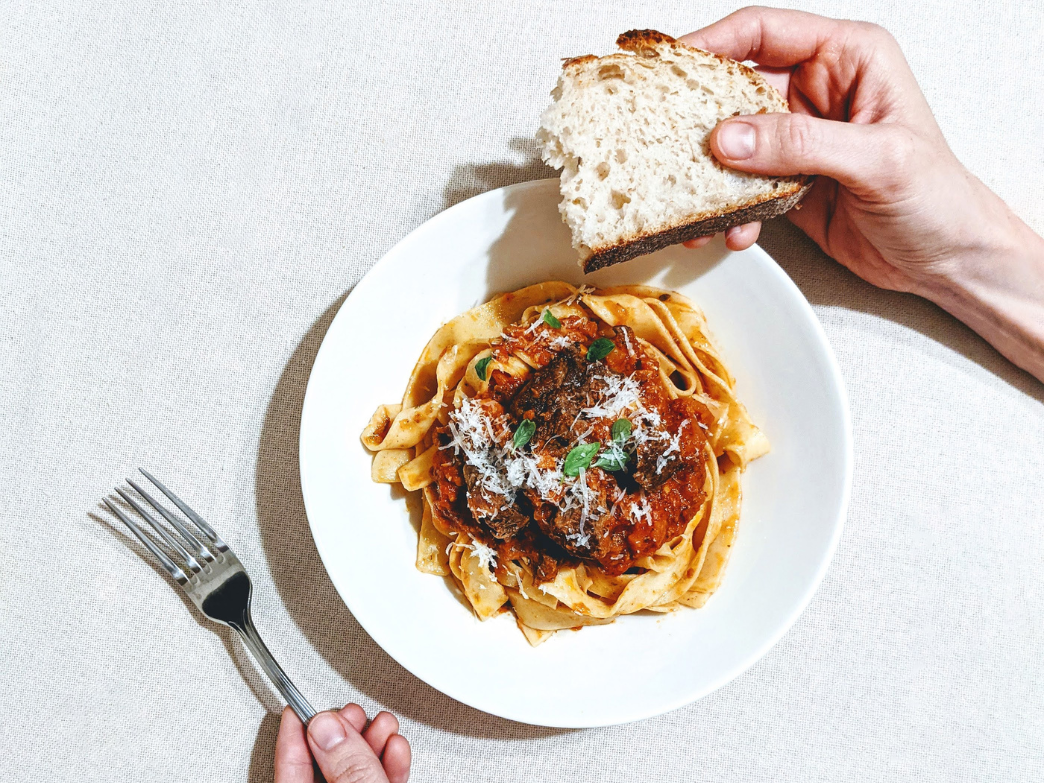 A plate of pasta with sauce and a piece of bread held in a hand.