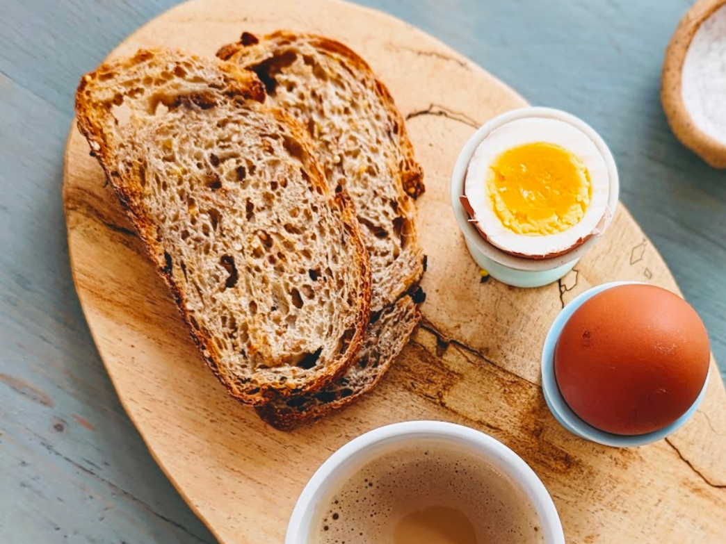 Two slices of multigrain bread with a soft-boiled egg, a whole egg, and a cup of coffee on a wooden board.