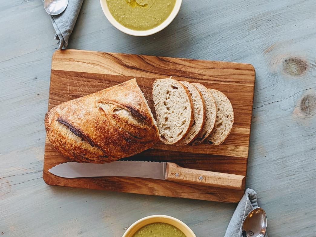 Sliced sourdough bread on a wooden cutting board with a knife and bowls of soup