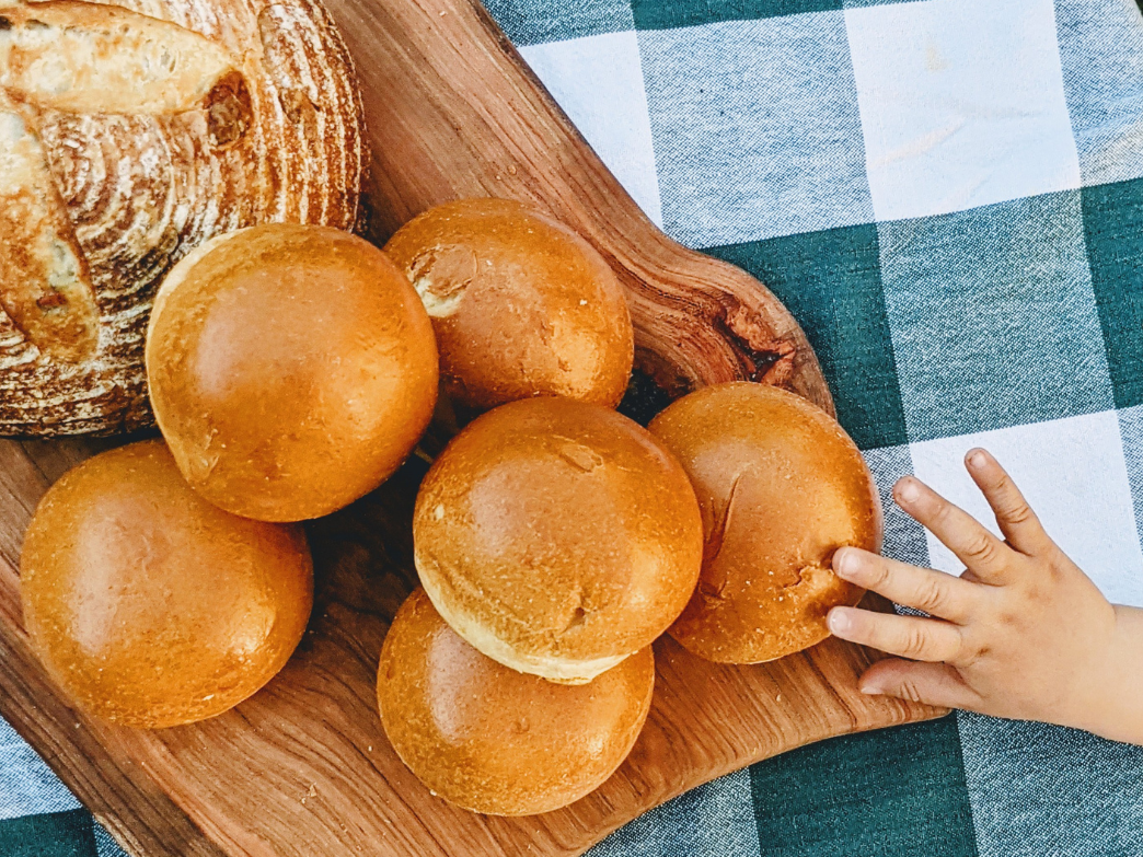 A wooden board with several golden brioche rolls and a hand reaching for one.