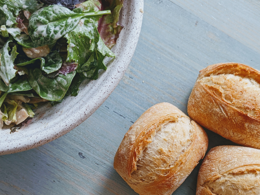 A bowl of salad next to three crusty bread rolls on a wooden table.