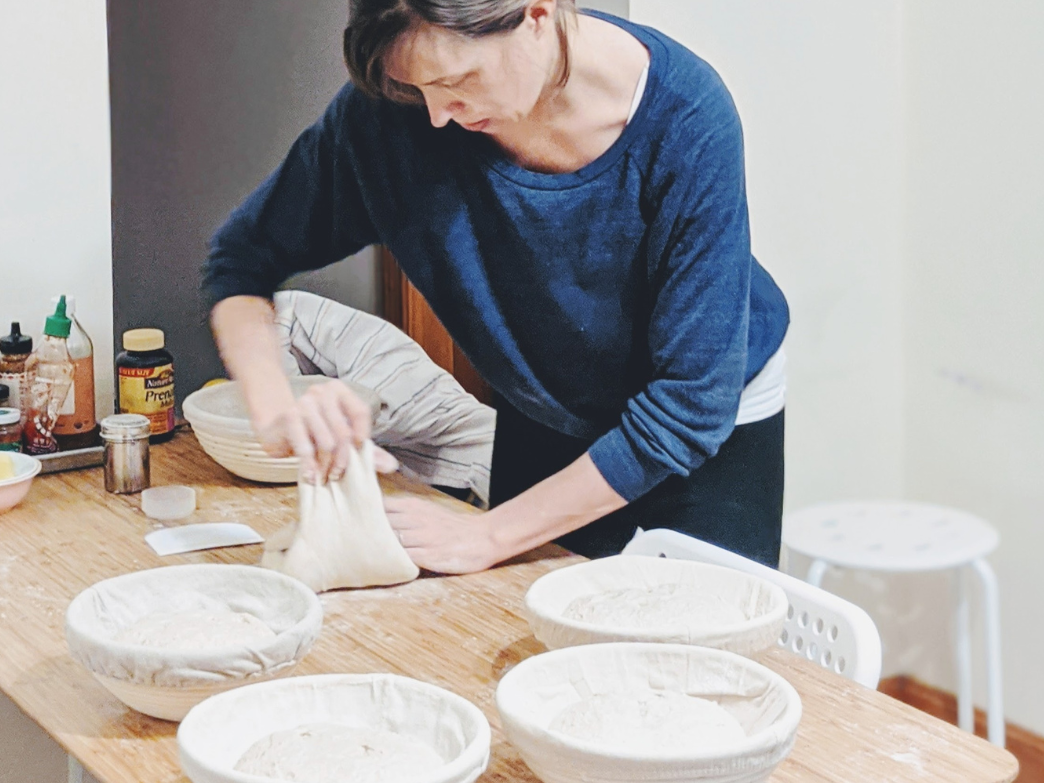 A person kneading dough on a wooden table with bowls of dough nearby.