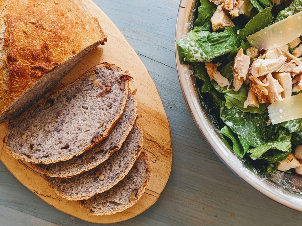 Sliced whole grain bread on a wooden board next to a salad with chicken and greens.