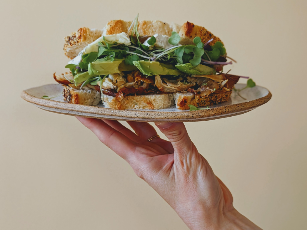 A hand holding a sandwich filled with greens, avocado, and other ingredients on a plate.